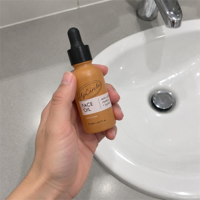 A hand holding UpCircle Organic Face Oil in its amber 30ml dropper bottle beside a washbasin